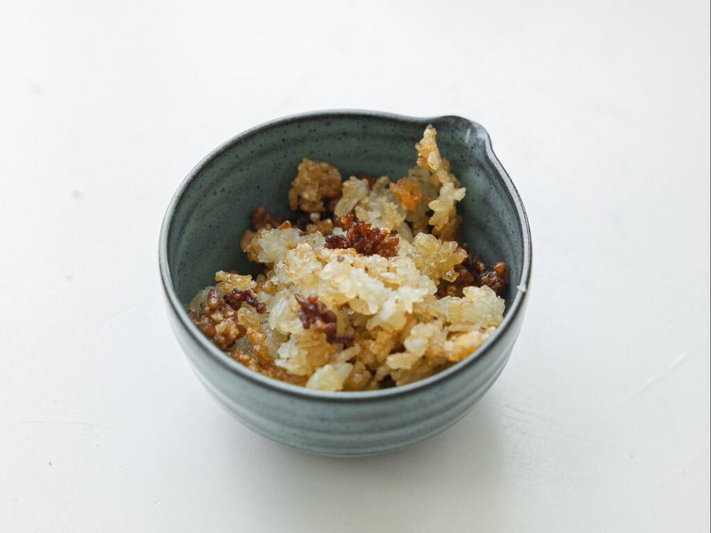 A small gray bowl filled with a mixture of cooked, crispy, golden-brown and white rice pieces, placed on a white surface.